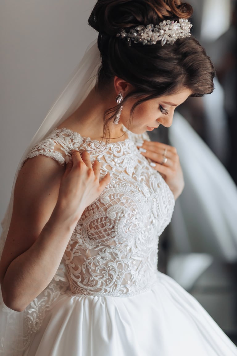 A woman in a wedding dress standing in front of a mirror adjusting her dress with veil and earrings