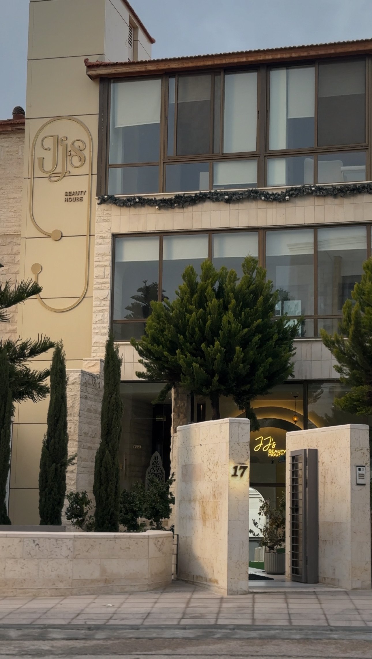 Modern multi-story building with cream facade, brown-framed windows decorated with garland, cypress plants flanking the entrance gate with house number 17
