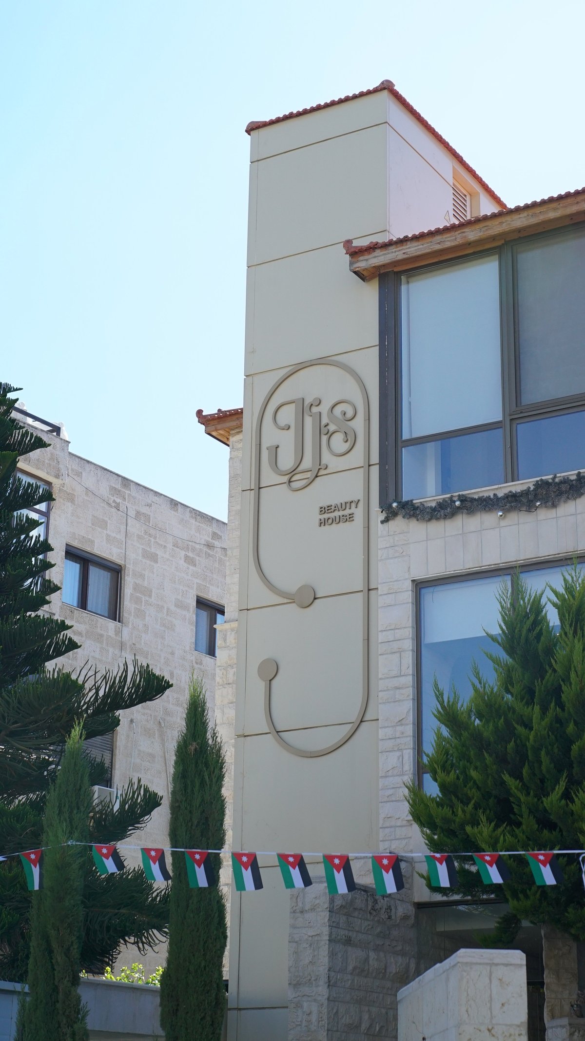 Modern cream-colored beauty house building with JB logo, red tile roof, large windows, and colorful flag bunting at base